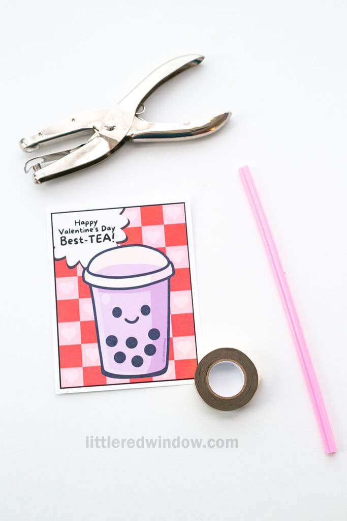 A Valentine’s card with a cartoon bubble tea and the message “Happy Valentine’s Day Best-TEA!” lies on a white surface next to a hole punch, a pink straw, and a roll of tape.
