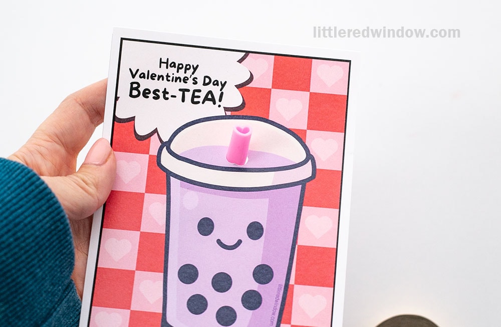 A hand holds a Valentine's Day card featuring a smiling bubble tea with a real pink heart straw. The card reads,