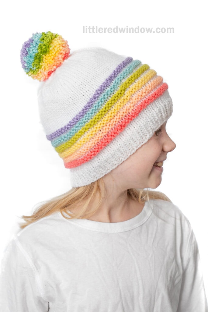 Young child wearing a white knit hat with a rainbow-striped band and a cheerful rainbow pom pom on top, smiling and looking to the side, dressed in a crisp white shirt.