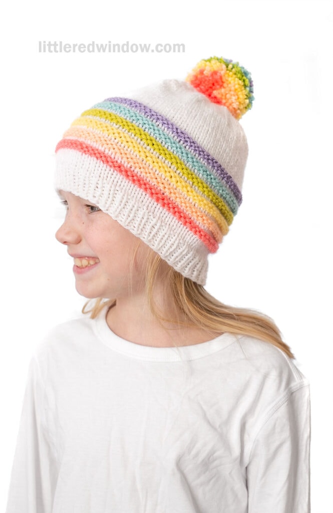 A smiling child wearing a white knit hat with rainbow pom pom and stripes, dressed in a plain white shirt, faces left.