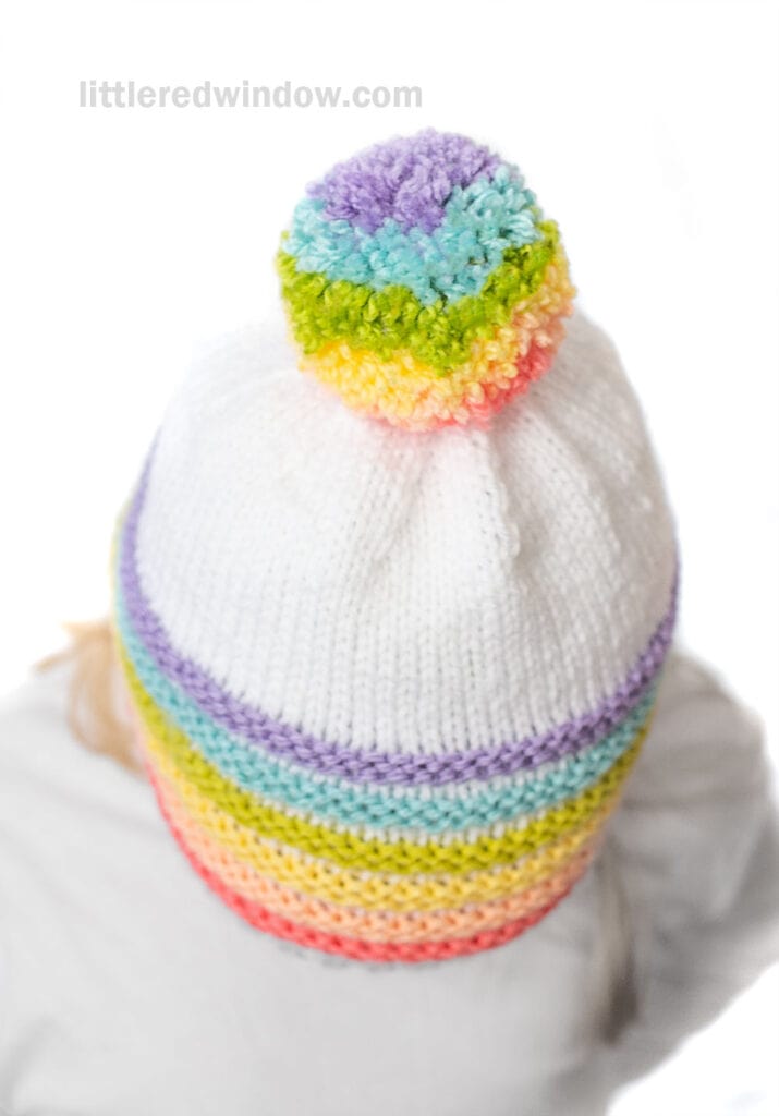 A child in a white knit hat with a rainbow pom pom and matching pastel stripes around the base, viewed from above on a white background.