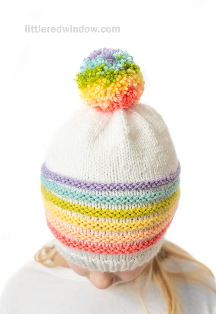 Child wearing a white knitted hat with a rainbow pom pom on top and colorful pastel stripes. The background is white, and the child’s face is mostly turned away.