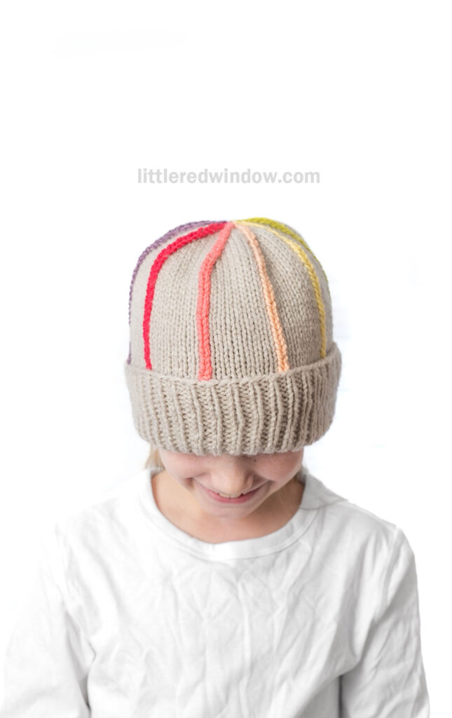 A child wearing a beige knit hat with colorful vertical stripes and a folded brim, looking down, on a white background.