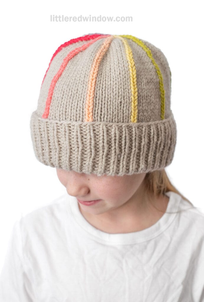A child wearing a beige knitted hat with colorful vertical stripes in red, orange, yellow, and pink, looking down with the hat covering their eyebrows. The child is dressed in a white shirt.