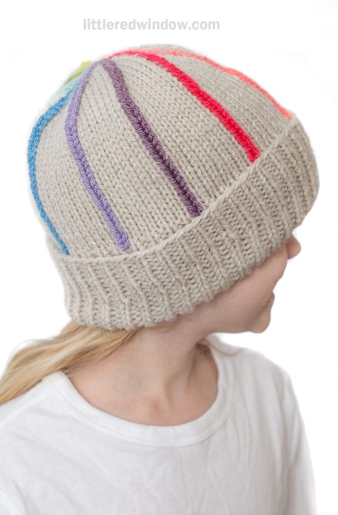 A child wearing a cream-colored knit hat with colorful, curved stripes and a folded ribbed brim, looking to the side. The child is dressed in a plain white shirt.