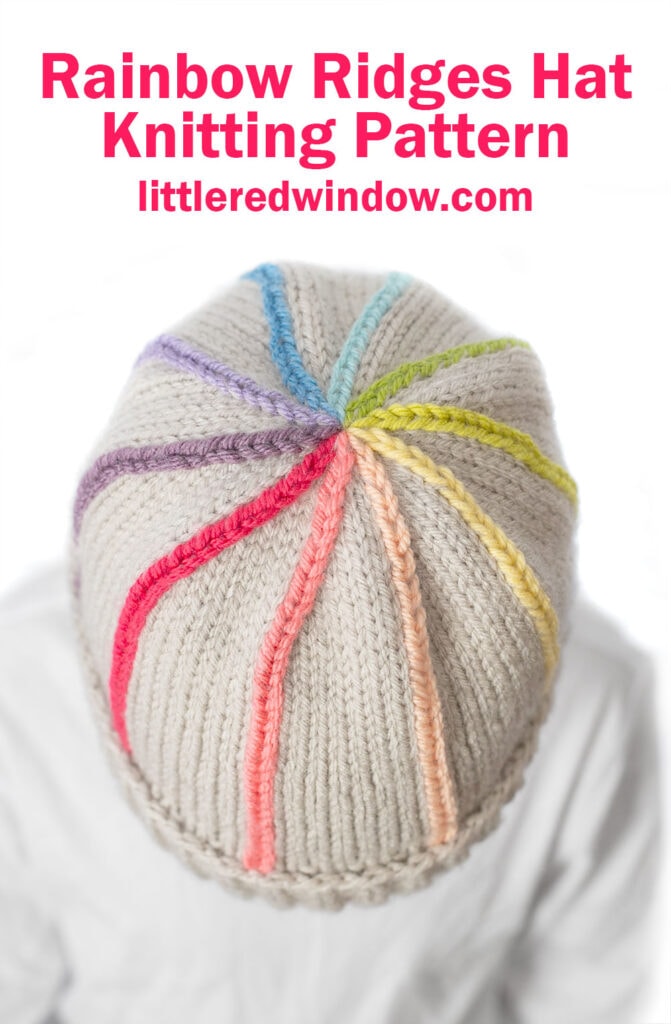 Top view of a cream-colored knit hat with colorful rainbow ridges radiating from the center, worn by a person in a white shirt. Pink text above reads: Rainbow Ridges Hat Knitting Pattern littleredwindow.com.