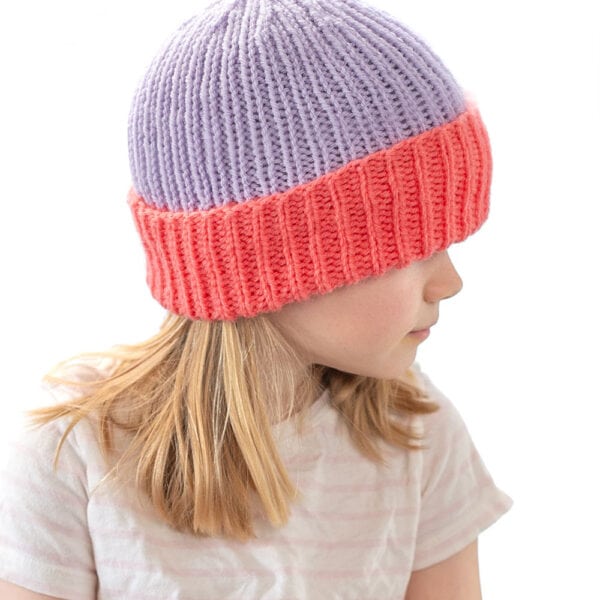 A young girl with blonde hair wears a colorblock folded brim hat featuring a purple top and bright pink brim. She looks to the side, and text above reads, “Colorblock Brim Hat Knitting Pattern littleredwindow.com.”.