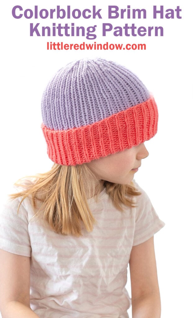 A young girl with blonde hair wears a colorblock folded brim hat featuring a purple top and bright pink brim. She looks to the side, and text above reads, “Colorblock Brim Hat Knitting Pattern littleredwindow.com.”.