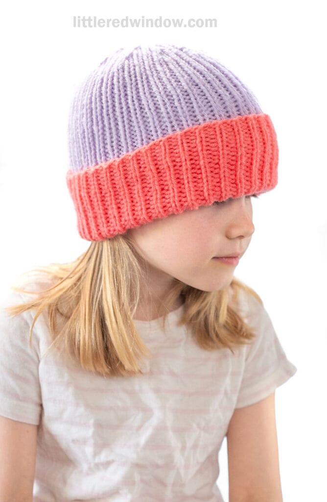 A young girl with blond hair, wearing a light purple and bright coral folded brim hat and a pale striped t-shirt, faces sideways against a white background. The beanie covers her eyebrows.