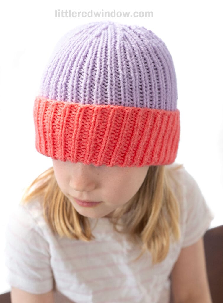 A child wearing a lilac and coral folded brim hat with a large brim looks down, the hat partially covering their eyes. The child is dressed in a light-striped shirt and has light brown hair.