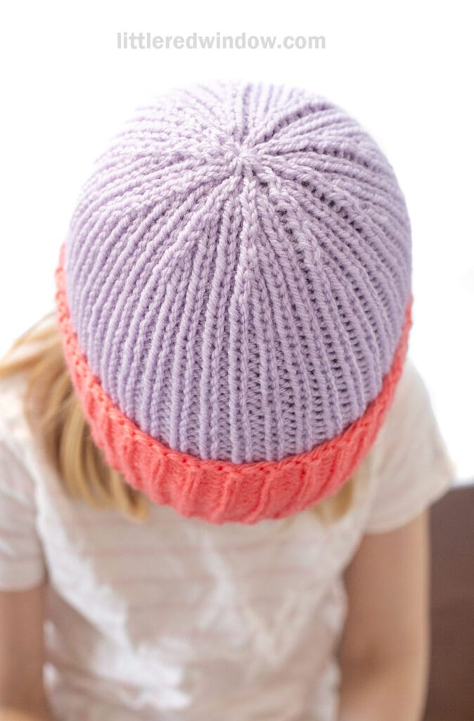 A child wearing a folded brim hat with a lavender top and coral ribbed brim looks down. The text