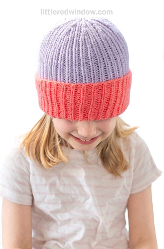 A young girl smiles while wearing a pink and purple folded brim hat pulled down over her eyes. She has light brown hair and is dressed in a light-colored, short-sleeve shirt against a plain white background.