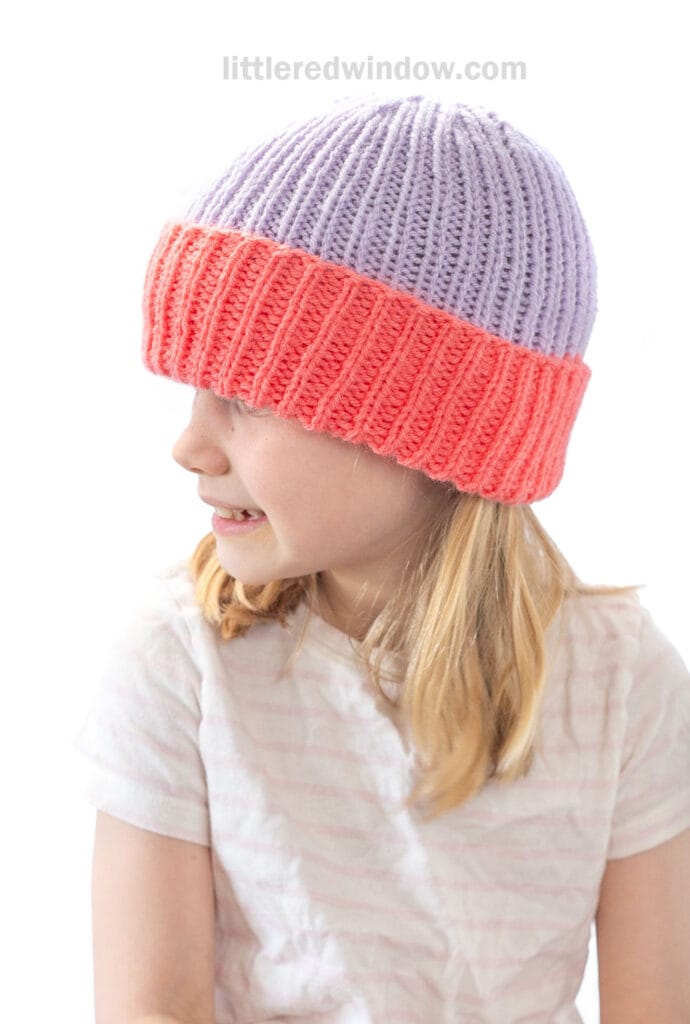 A young child with blonde hair wears a knit folded brim hat with a purple top and wide pink edge, looking to the side and smiling. The child wears a light-colored shirt.