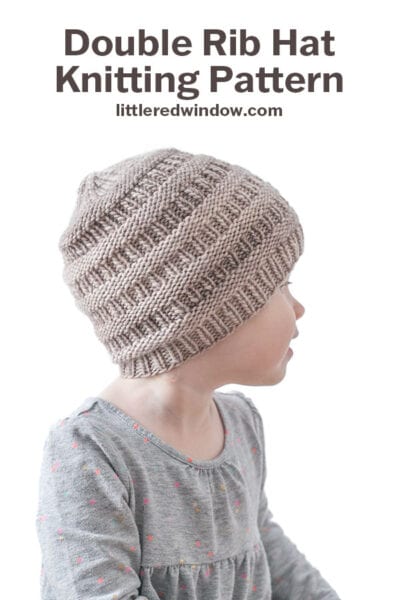 A young child wearing a gray Rib Hat with a double rib pattern looks to the side. Text above reads: “Double Rib Hat Knitting Pattern littleredwindow.com.” The child is in a gray shirt with small colored dots.