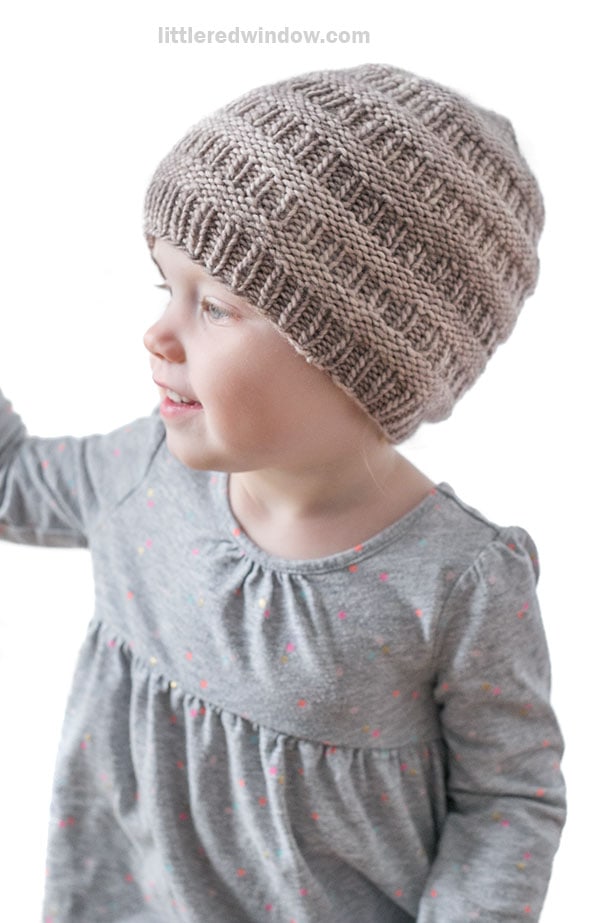 Young child wearing a light brown rib hat and a gray long-sleeve dress with small colorful dots, looking to the side and smiling against a white background.