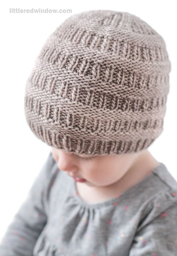 A young child wearing a knitted beige rib hat with a textured pattern, looking down while dressed in a gray shirt with small colorful dots.