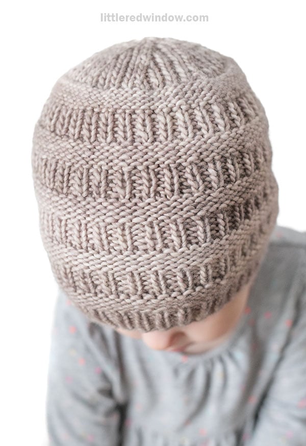 A child wearing a taupe-colored rib hat with a textured, ridged pattern looks downward. The child is dressed in a light grey shirt adorned with small pink and blue dots.