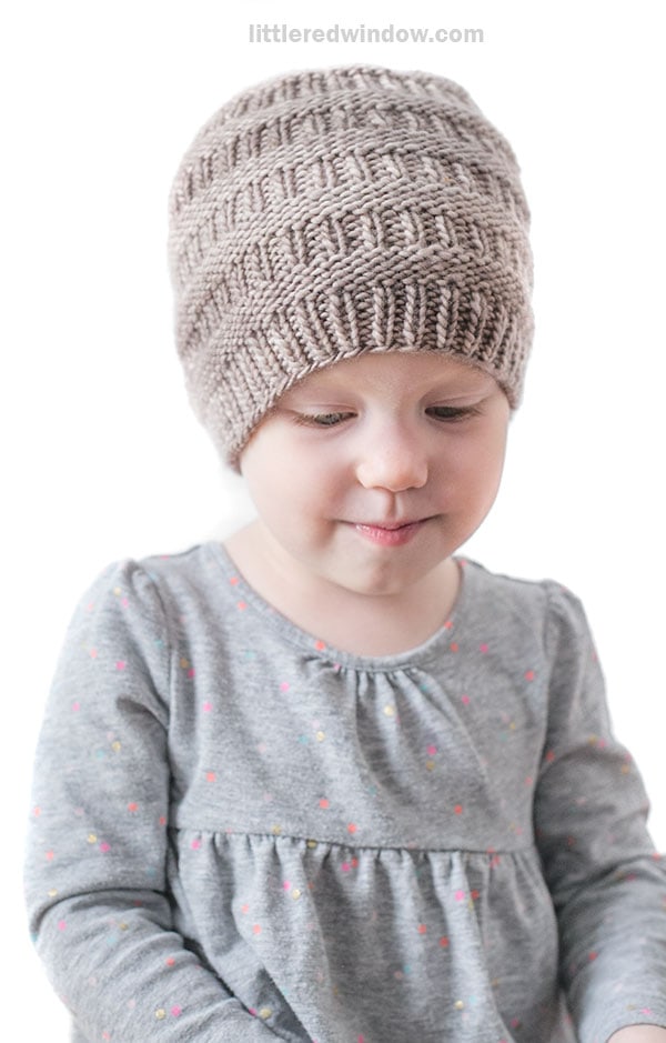 A young child wearing a textured knit Rib Hat in beige and a gray long-sleeve shirt with small colorful dots, looking down with a slight smile against a plain white background.