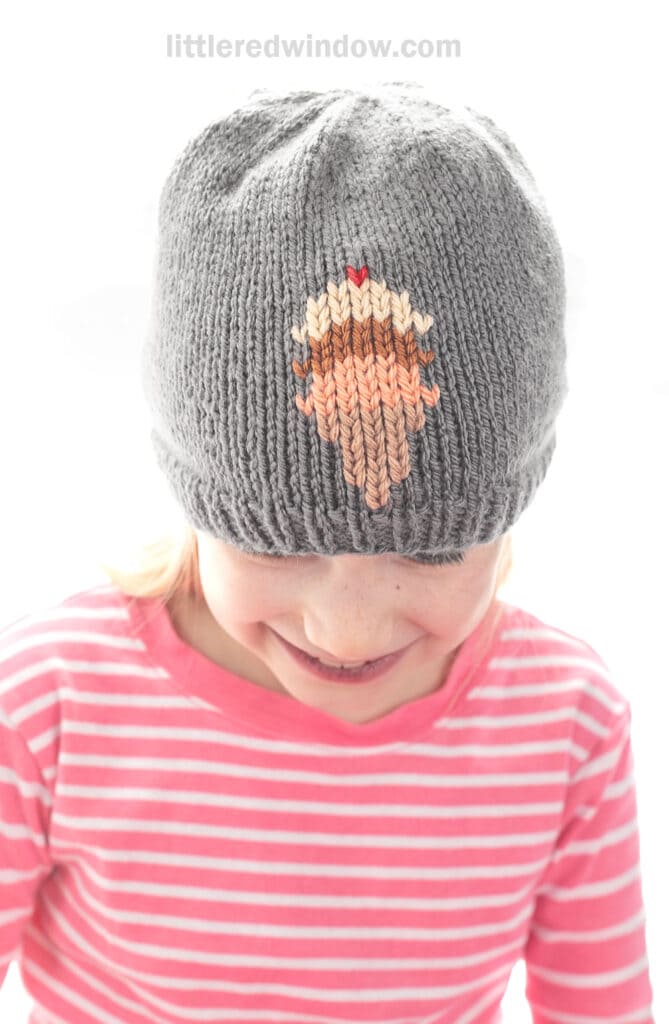 A child wearing a gray knit hat with a cupcake design, reminiscent of an ice cream cone hat, smiles while looking down. The child is dressed in a pink and white striped shirt.