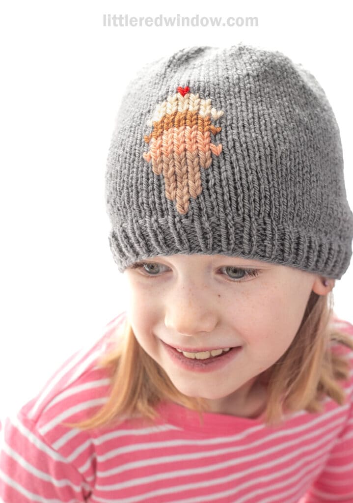 A smiling child wears an ice cream cone hat with a gray knit design, paired with a pink and white striped shirt.