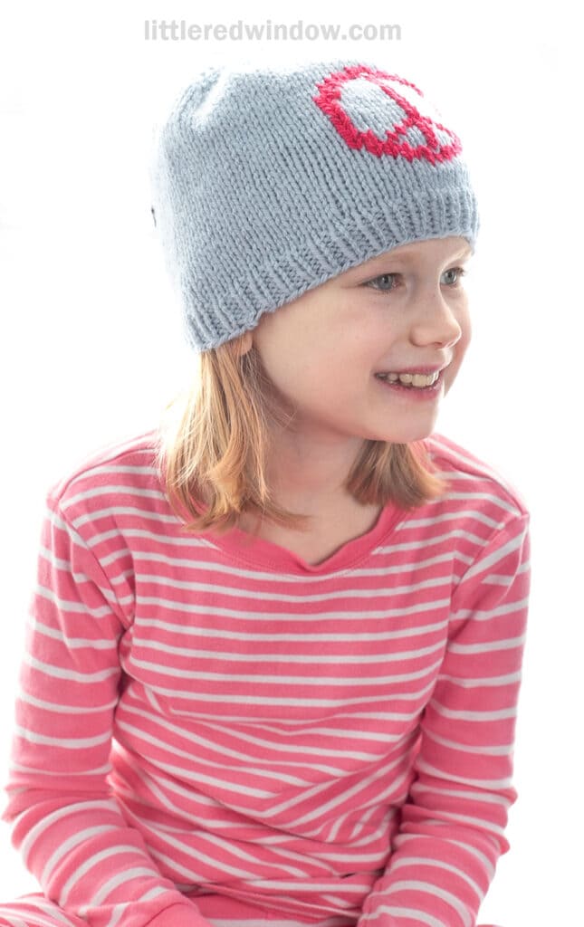 A young girl with blonde hair wears a light blue knit hat with a pink and white skull design and a pink and white striped long-sleeve shirt. She is smiling and looking slightly to the side.