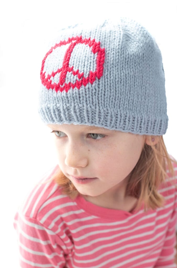 A young child wearing a light gray knit hat with a pink peace symbol and a pink-striped shirt, looking slightly downward against a white background.