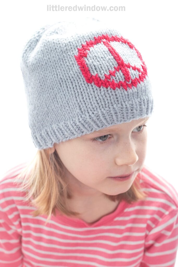 Young child wearing a light gray knitted hat with a red peace sign design, paired with a pink and white striped shirt, looking slightly to the side.