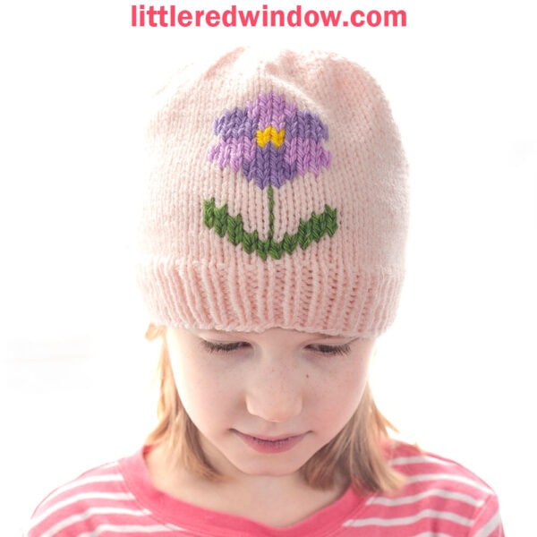 Young girl wearing a light pink flower hat with a purple flower and green leaves design, looking down. Text above reads: