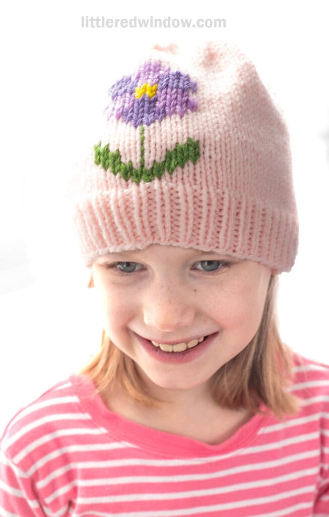 Smiling young girl wearing a light pink flower hat decorated with a purple blossom and green leaves, paired with a pink and white striped shirt.