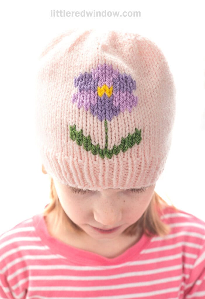 A child wearing a charming flower hat—a light pink knit hat with a purple and yellow flower and green stem design—looks down, dressed in a pink and white striped shirt.