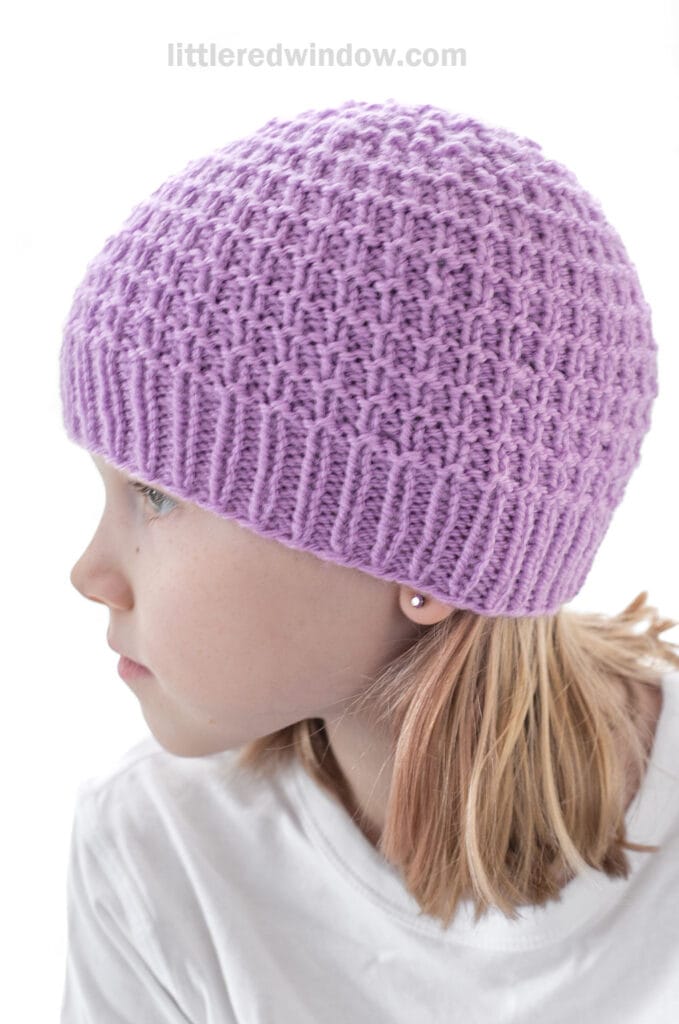 A young girl with blonde hair, wearing a textured lavender knit hat and a white shirt, looks to the side against a white background.