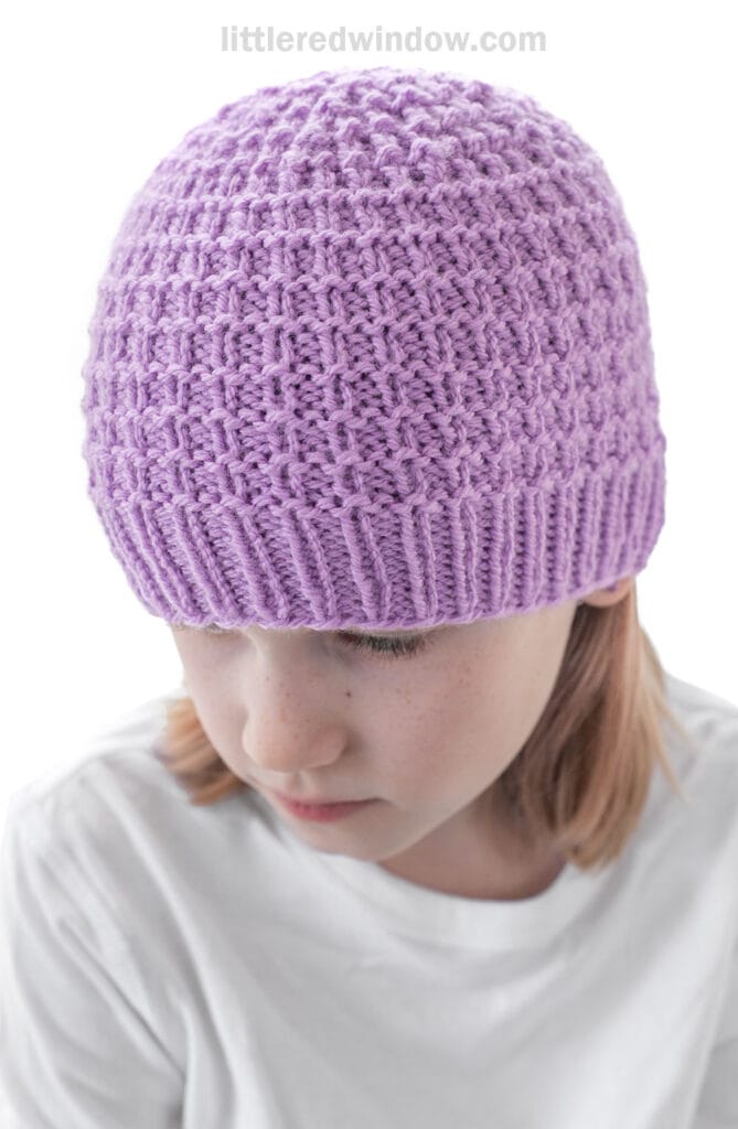 A child wearing a light purple, textured knit hat and a white shirt looks downward against a plain white background.