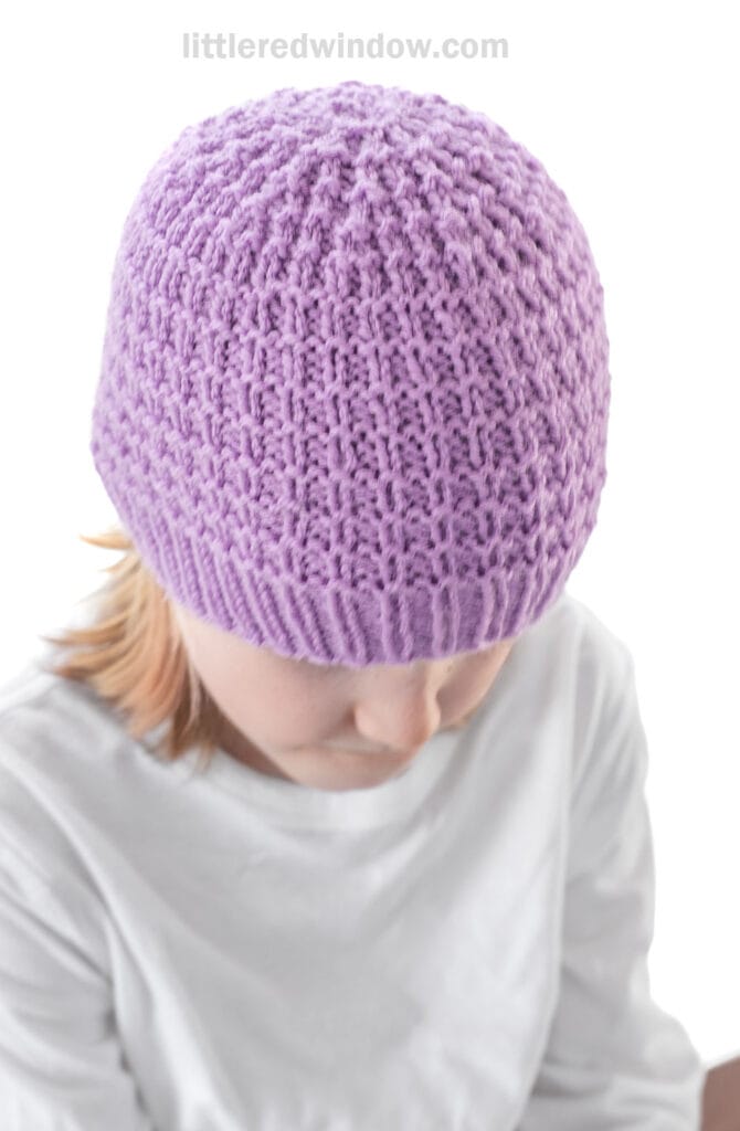 Person wearing a textured, lavender purple knit hat and a white shirt, looking downward against a white background.