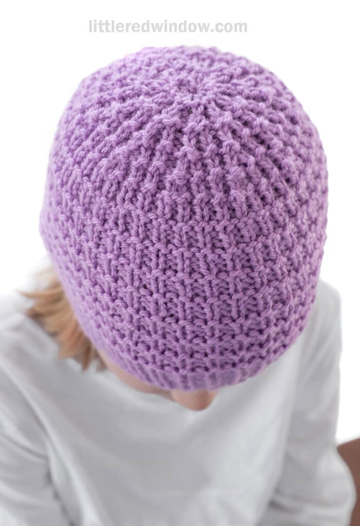 A person wearing a light purple, textured knit hat, viewed from above. The person is dressed in a white shirt and looking downward, with only the top of their head and shoulders visible.