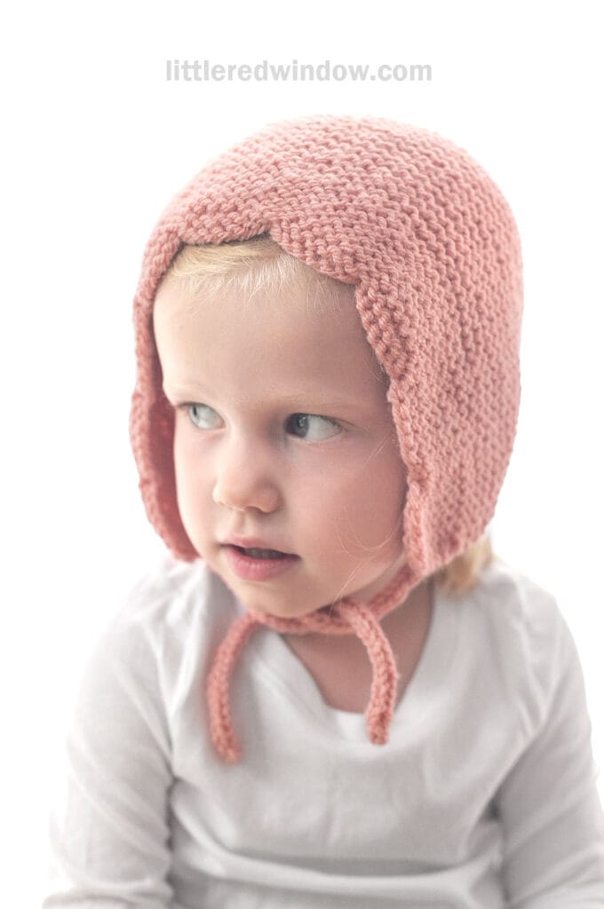 A young child wears a pink scalloped bonnet with ties under the chin, looking slightly to the side against a white background. The text