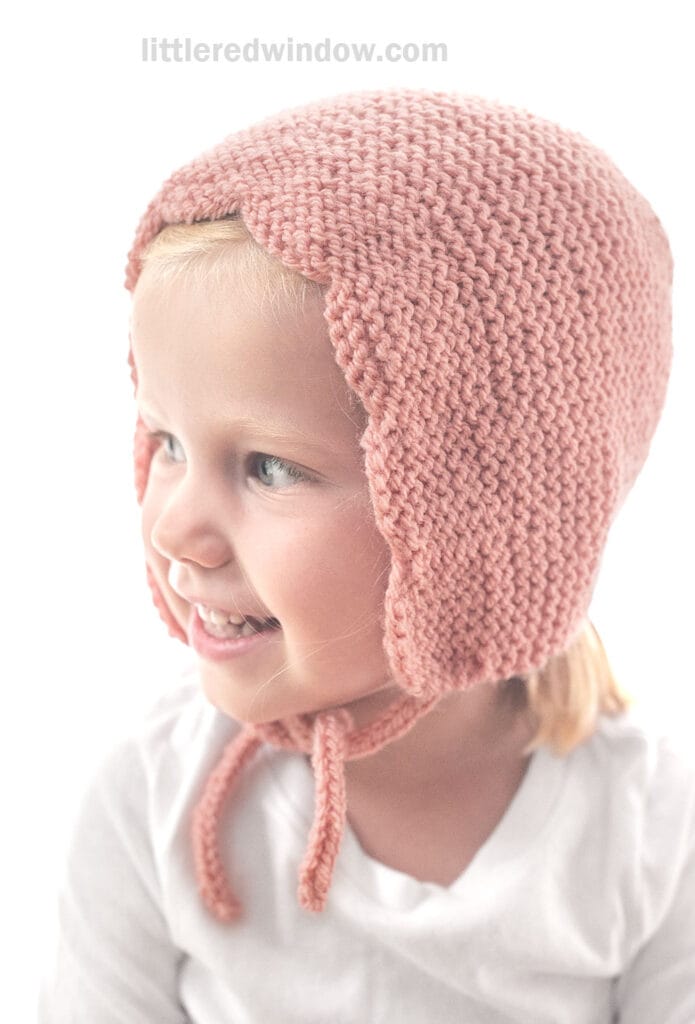 A young child wearing a pink scalloped bonnet with ties, smiling and looking to the side. The background is white and the child is dressed in a white shirt.