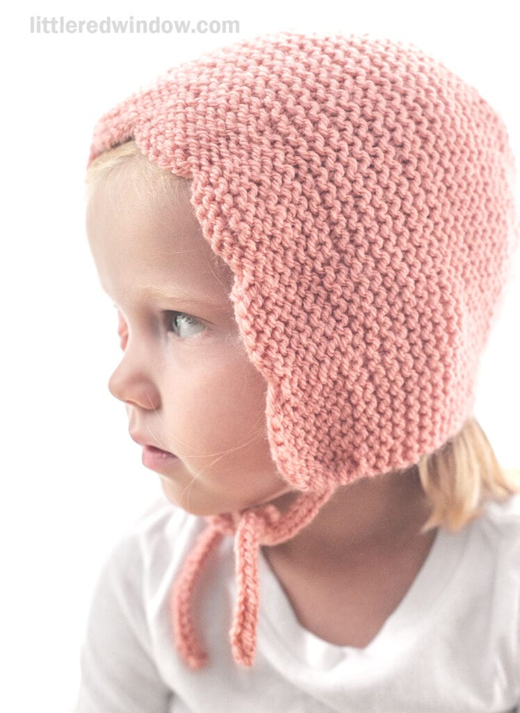 A young child wearing a hand-knitted, pink scalloped bonnet with tied straps, looking to the left, against a plain white background.