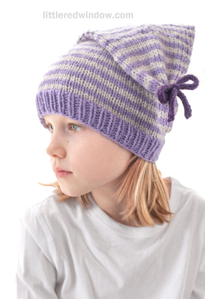 A young child with blond hair wears a white shirt and a purple-and-white striped tied stocking cap with a pointed top and bow, looking off to the left against a white background.