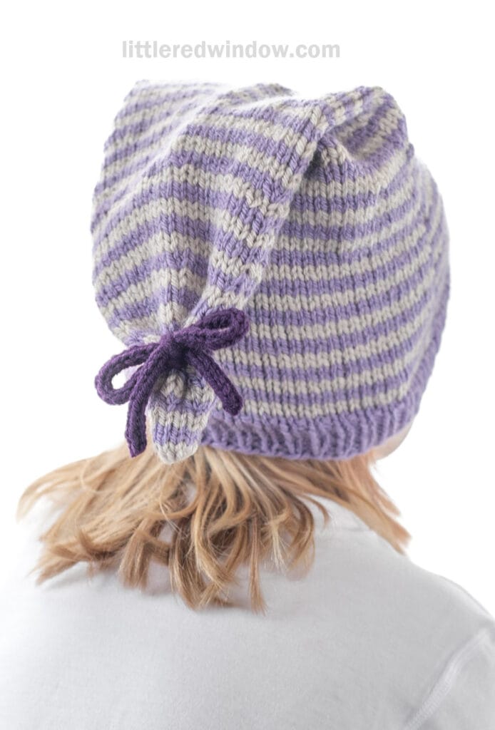 A person with light brown hair is wearing a tied stocking cap—knitted purple and gray with stripes and a pointy end tied in a bow at the back. The hat has ribbed edging and is shown from behind.