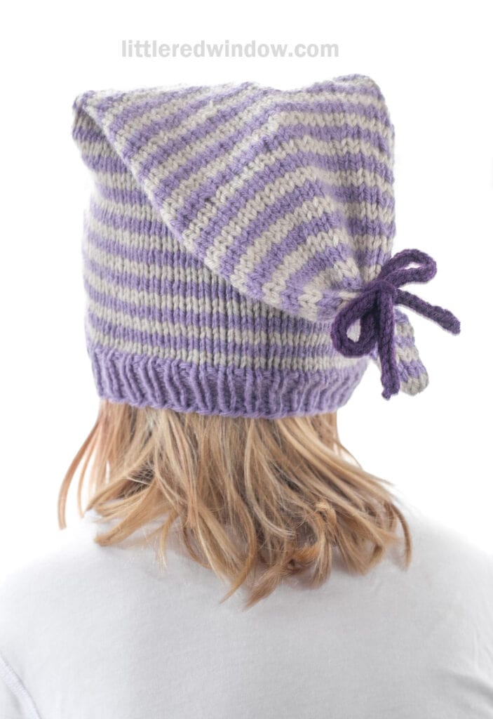 A person with light brown hair wears a hand-knit, purple and white striped tied stocking cap with a bow on the side. The photo is taken from behind, and the person is dressed in a white shirt.