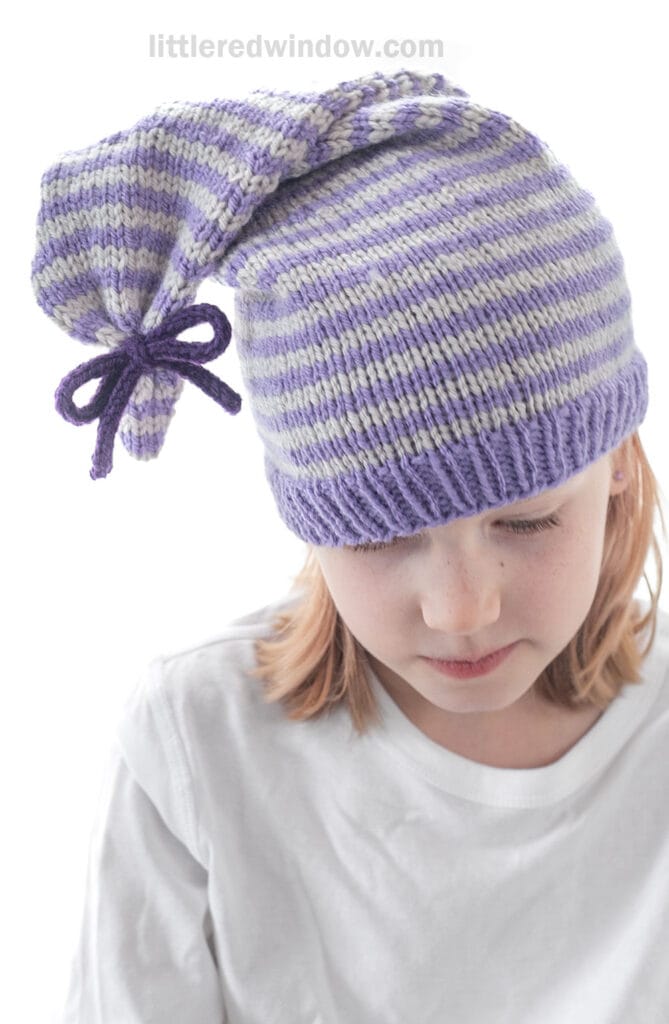 A child with light brown hair wears a white shirt and a purple and white striped tied stocking cap with a pointed, floppy top in a bow. The child looks downward against a clean, white background.
