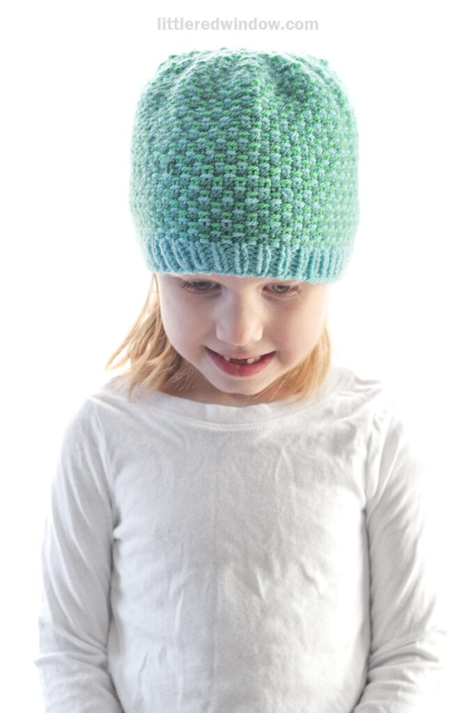 A young child wearing a light blue and green linen stitch hat and a plain white long-sleeve shirt looks down and smiles against a white background.