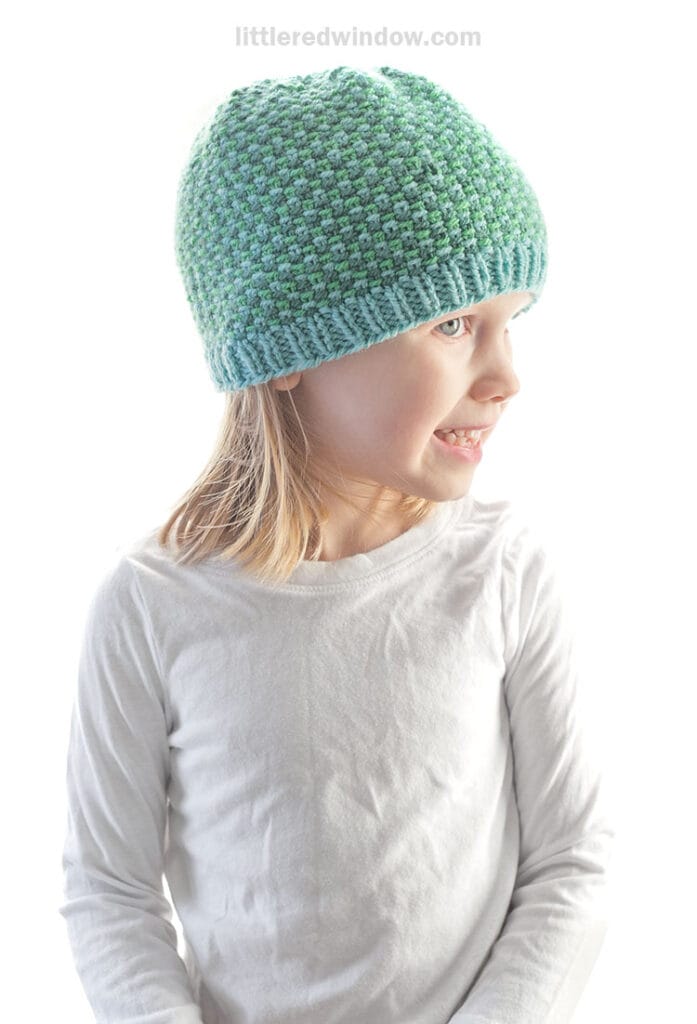 A young child with long blonde hair wears a light blue and green linen stitch hat and a white long-sleeve shirt, smiling and looking to the side against a white background.