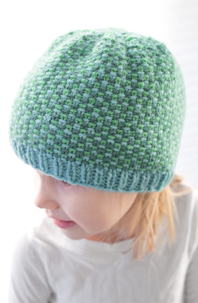 A young child wearing a textured, light blue and green linen stitch hat looks down and to the side, with blonde hair peeking from under the beanie and a white shirt. The background is softly blurred.