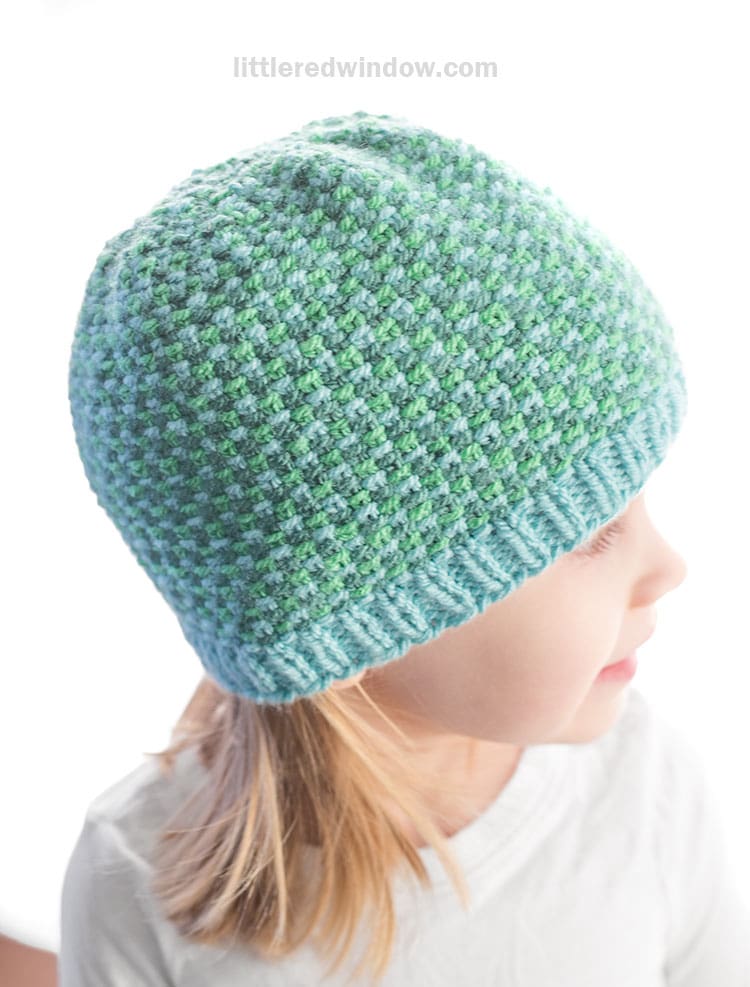 A young child wearing a blue and green linen stitch hat, looking to the right, with light brown hair peeking out from under the hat and a white shirt.