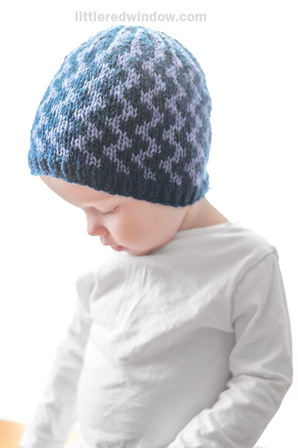 A young child wearing a white shirt and a blue patterned knit hat with wavy lines looks down, standing against a bright white background.