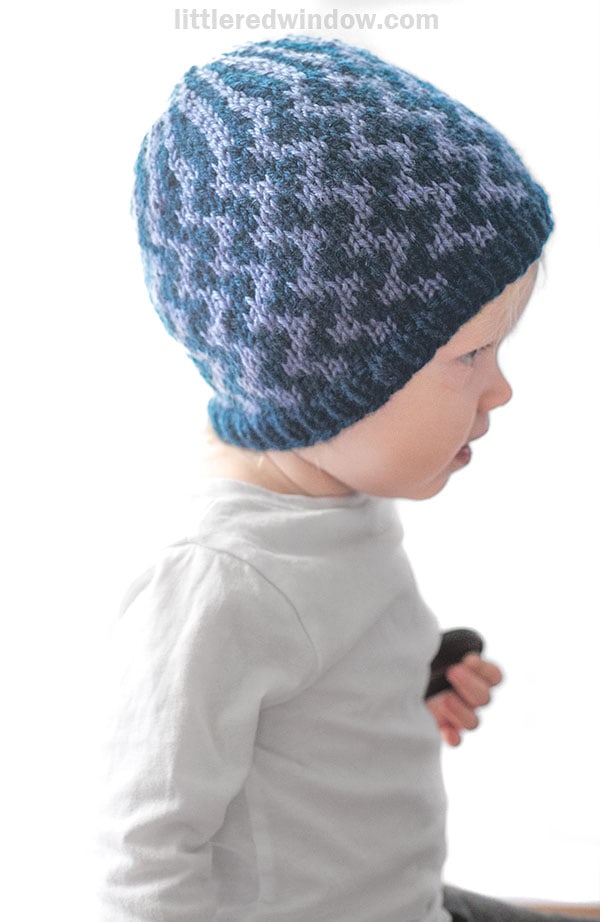 A young child wearing a white shirt and a blue and purple patterned knit hat featuring wavy lines sits in profile against a white background.