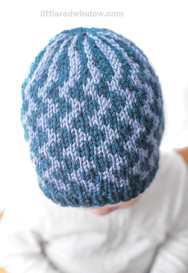 Top view of a child wearing a knitted blue and light purple hat with a geometric pattern featuring wavy lines, sitting against a white background.