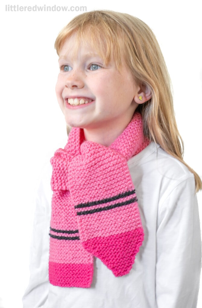 Smiling young girl with blonde hair wearing a white shirt and a playful crayon scarf with dark stripes, standing against a white background.