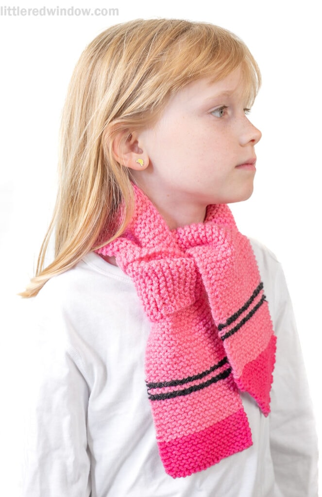 A young girl with blonde hair and gold heart earrings wears a white shirt and a pink crayon scarf with black stripes, looking to the side against a white background.